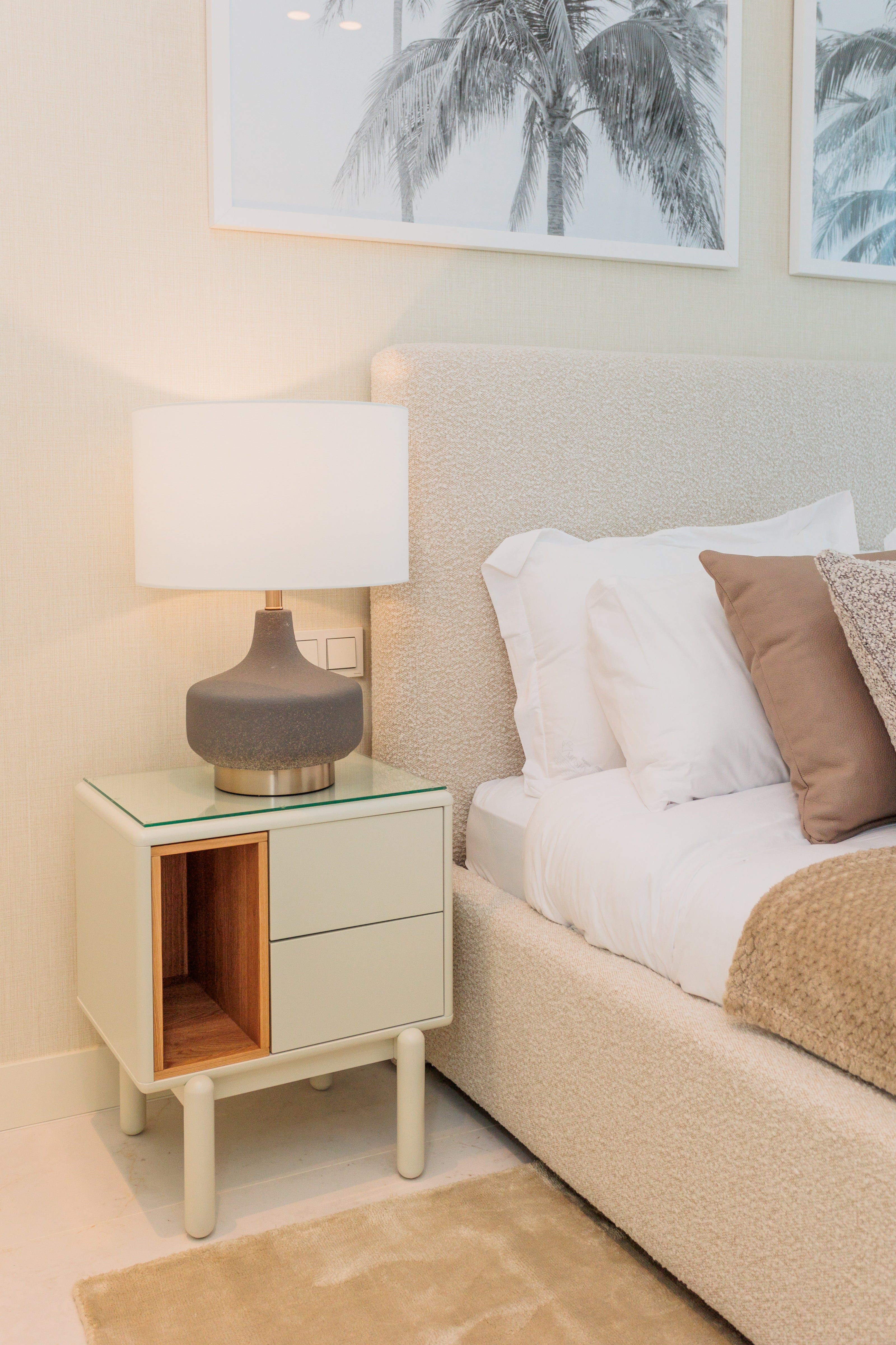 luxury soft interior design for bedroom with white bedside table, beige cream bed and headboard, greyish brown table lamp, palm tree artwork and brown rug available in benalmadena, marbella, malaga, costa del sol.