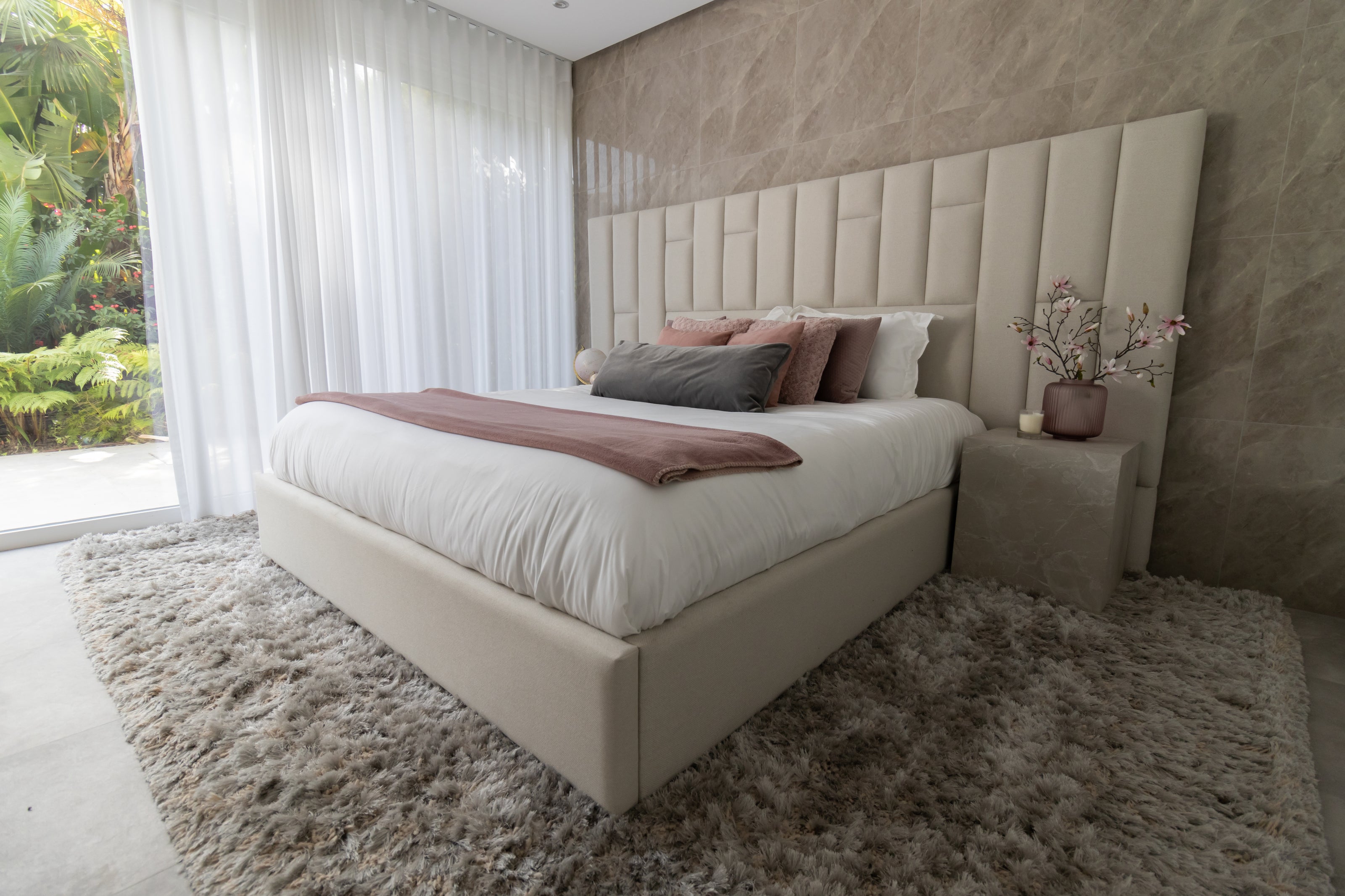 elegant bedroom for real estate property home staging interior design which includes bed with beautiful headboard, marble bedside table, shaggy beige brown rug, custom made white voiles and pink pillows.